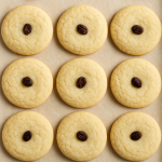 A high-resolution photograph showcases nine freshly baked sugar cookies, each with a single raisin centered on top, arranged neatly on a parchment-lined baking sheet.