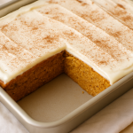 A freshly baked rectangular pumpkin cake topped with a thick layer of cream cheese frosting and sprinkled with cinnamon, sitting on a cooling rack.