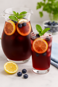 Glass pitcher and glass of blueberry iced tea with lemon slices, fresh blueberries, and mint on a marble surface