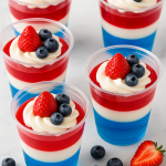 Five patriotic-themed layered Jello parfaits in clear cups, topped with whipped cream, blueberries, and strawberries on a light wooden surface