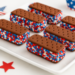 Six vanilla ice cream sandwiches rolled in red, white, and blue sprinkles, displayed on a festive white tray for Fourth of July