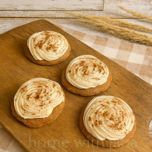 Four frosted pumpkin spice cookies topped with cream cheese frosting and a sprinkle of cinnamon, displayed on a wooden board.