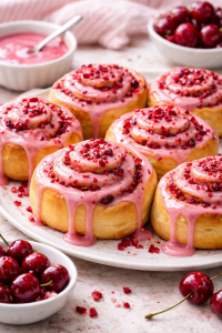 Close-up of freshly baked cherry sweet rolls topped with glossy pink glaze and bits of cherry, arranged on a white plate with bowls of glaze and cherries in the background