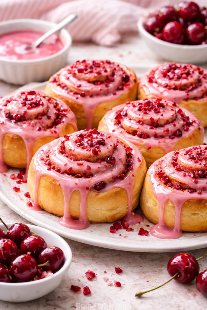 Close-up of freshly baked cherry sweet rolls topped with glossy pink glaze and bits of cherry, arranged on a white plate with bowls of glaze and cherries in the background