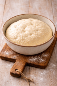 Homemade yeast dough in a ceramic bowl resting on a wooden bread board