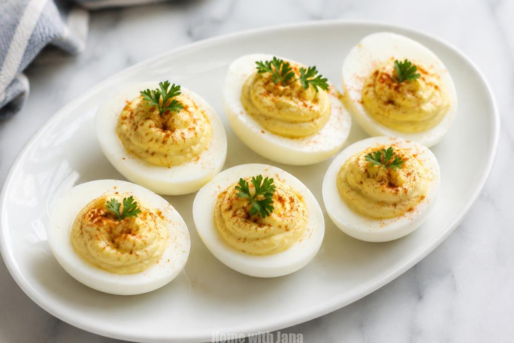 Classic deviled eggs arranged on a white oval platter with creamy pale yellow filling, sprinkled with paprika and topped with fresh parsley. Soft natural lighting and a clean background create a simple homemade appetizer presentation.