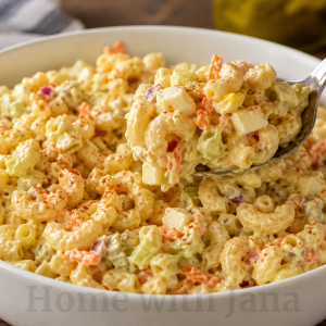 Creamy old-fashioned macaroni salad in a large white ceramic bowl with elbow macaroni, chopped hard-boiled eggs, celery, red onion, shredded carrots, and sweet pickle relish mixed in a pale yellow dressing. A large spoon lifts a hearty scoop of the salad, showing the chunky texture. Light paprika sprinkled on top. Rustic wooden table with warm natural window lighting.
