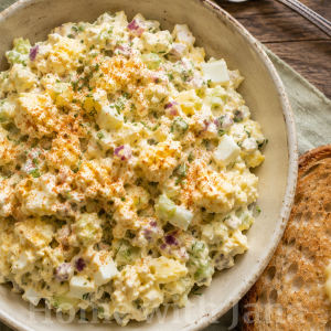 Overhead view of a rustic bowl of homemade egg salad made with chopped hard-boiled eggs, diced celery, red onion, fresh dill, and chives in a creamy dressing, lightly dusted with paprika. The bowl sits on a soft sage linen napkin beside a slice of toasted sourdough bread topped with melting butter. A vintage silver spoon, fresh dill sprigs, and an open cookbook rest on a weathered wooden table in warm natural light.